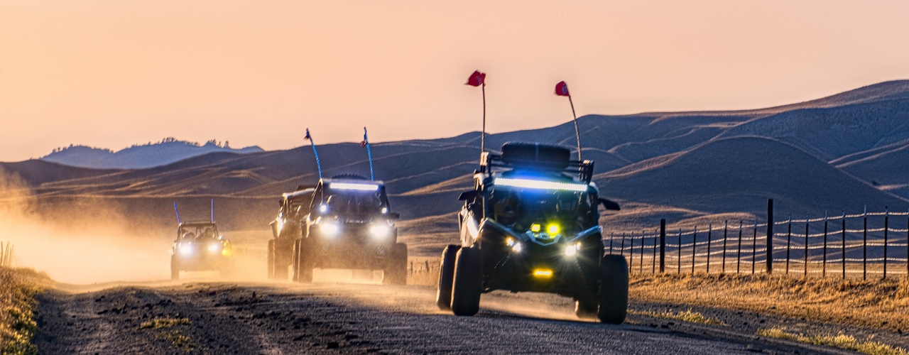 A line of four UTVs driving on a dirt road. There are large dunes of sand behind them in the distance.