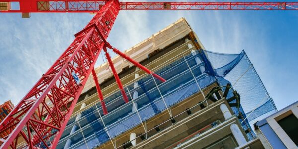 A ground-level view looking up at a large red construction crane near a high-rise building under construction.