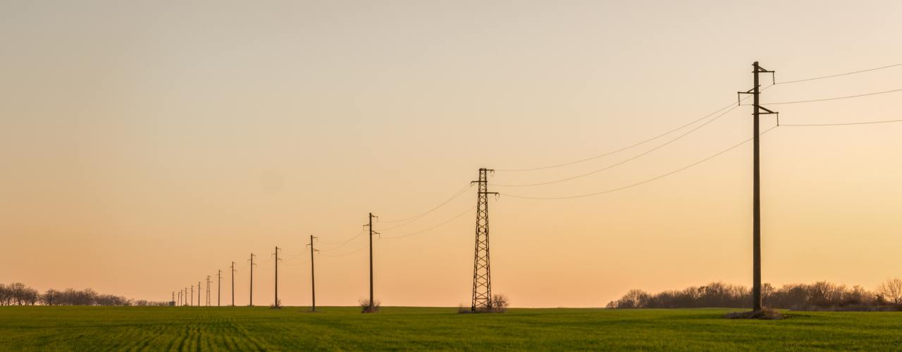 A line of electric poles running across an open field with green grass. The sun is setting behind the poles.