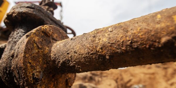 A close-up of a rusted industrial pipe in a dirt trench, with excavation equipment blurred in the background.