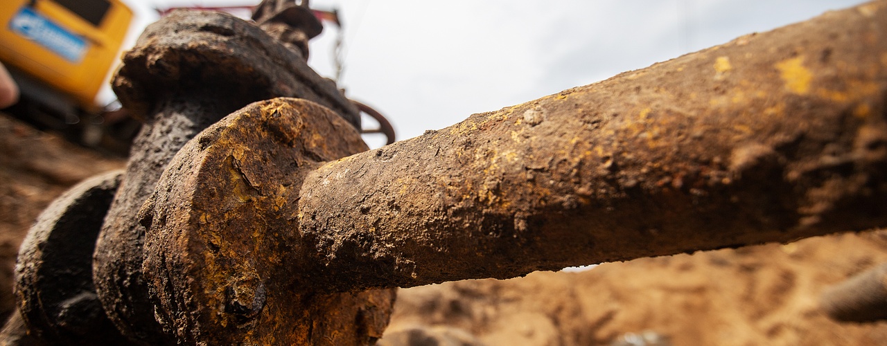 A close-up of a rusted industrial pipe in a dirt trench, with excavation equipment blurred in the background.