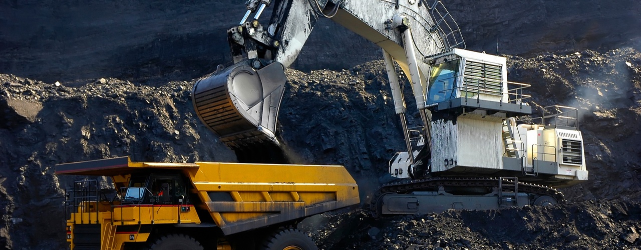 An operator in a white excavator loads up coal into a yellow bedding box as it works on a coal mining job site.