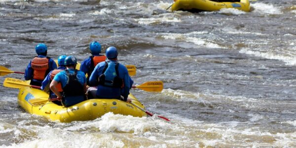 Groups of people wearing helmets and life vests raft down a fast-moving river through forested rapids.