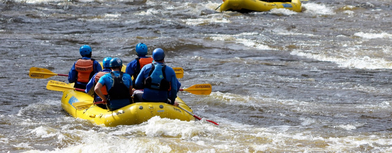 Groups of people wearing helmets and life vests raft down a fast-moving river through forested rapids.