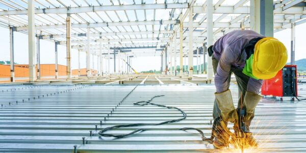 A construction worker bending over to weld steel panels at his feet. The steel building's frame arcs over him.