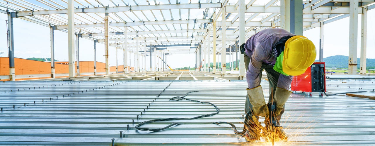 A construction worker bending over to weld steel panels at his feet. The steel building's frame arcs over him.