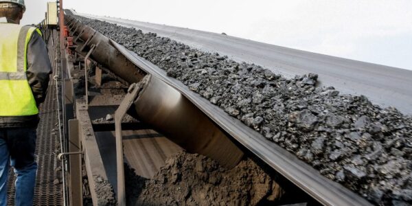 A mine worker walks up a ramp alongside a conveyor system that transports pieces of coal. The sky is clear.