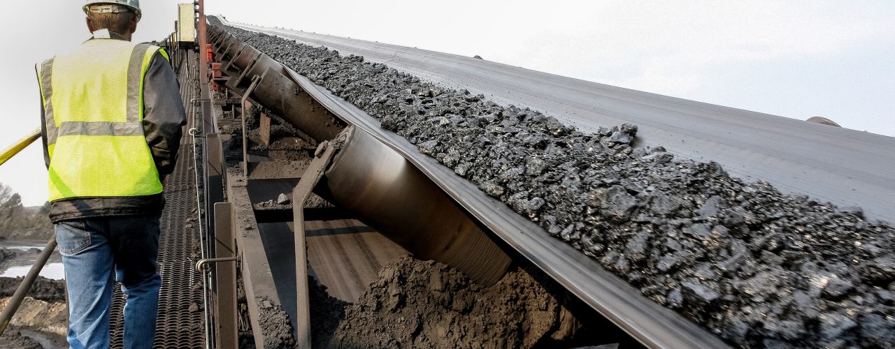 A mine worker walks up a ramp alongside a conveyor system that transports pieces of coal. The sky is clear.