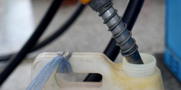 Fuel nozzle inserted into plastic container opening, with hose and equipment blurred in background of industrial setting.
