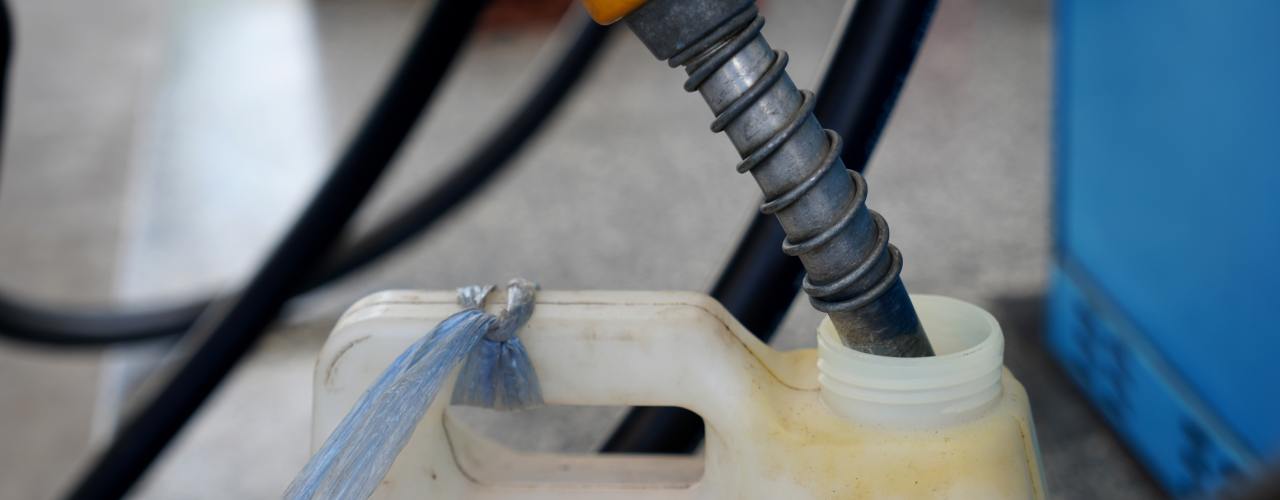 Fuel nozzle inserted into plastic container opening, with hose and equipment blurred in background of industrial setting.