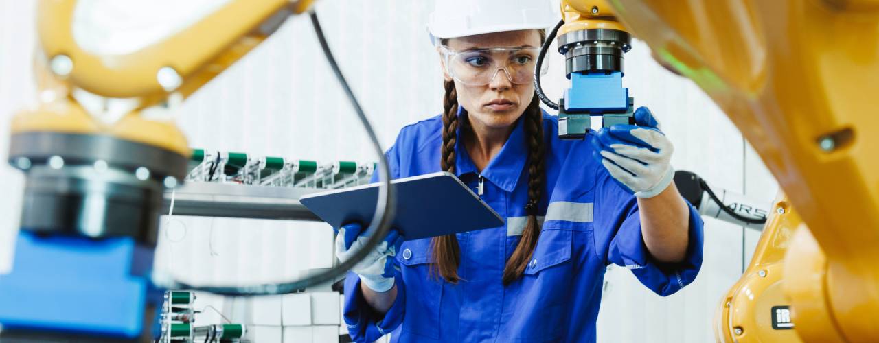 An engineer wearing safety glasses adjusting an industrial machine with one hand and holding a tablet in the other hand.