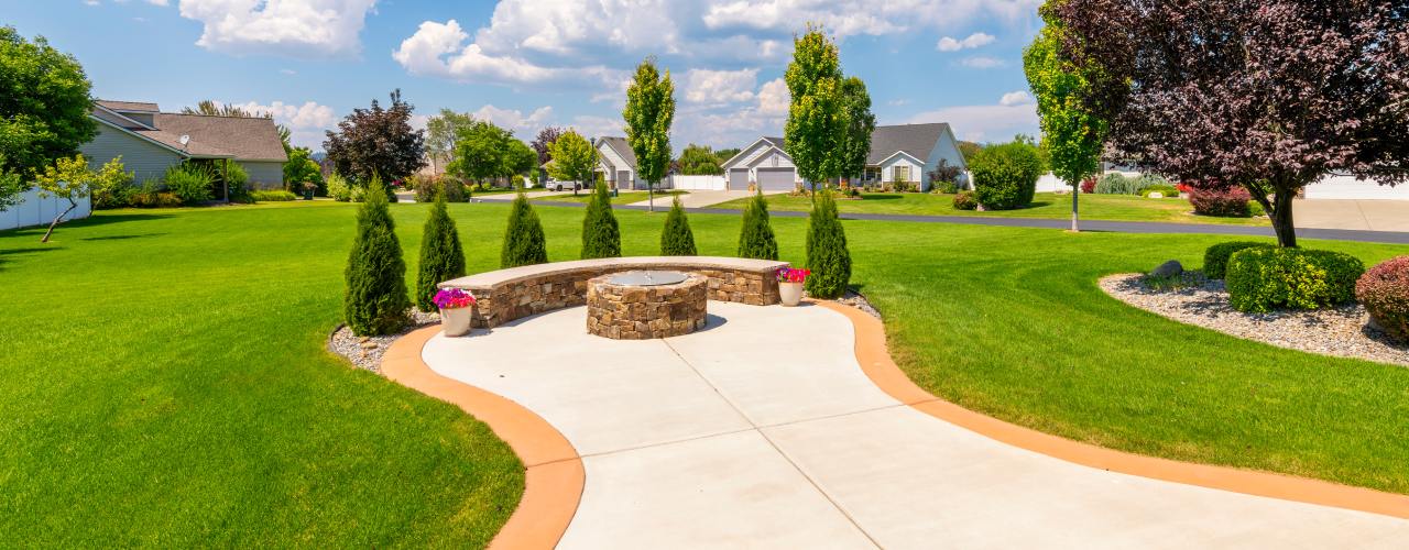 A backyard with a functional firepit in the center of a paved walkway. The bench seating is surrounded by mini trees.