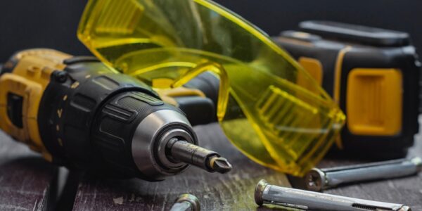 Cordless drill with bit, yellow safety glasses, tape measure, and metal anchors on worn wooden surface.