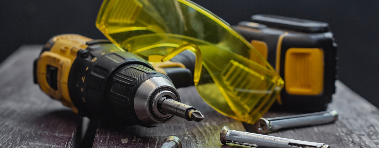 Cordless drill with bit, yellow safety glasses, tape measure, and metal anchors on worn wooden surface.
