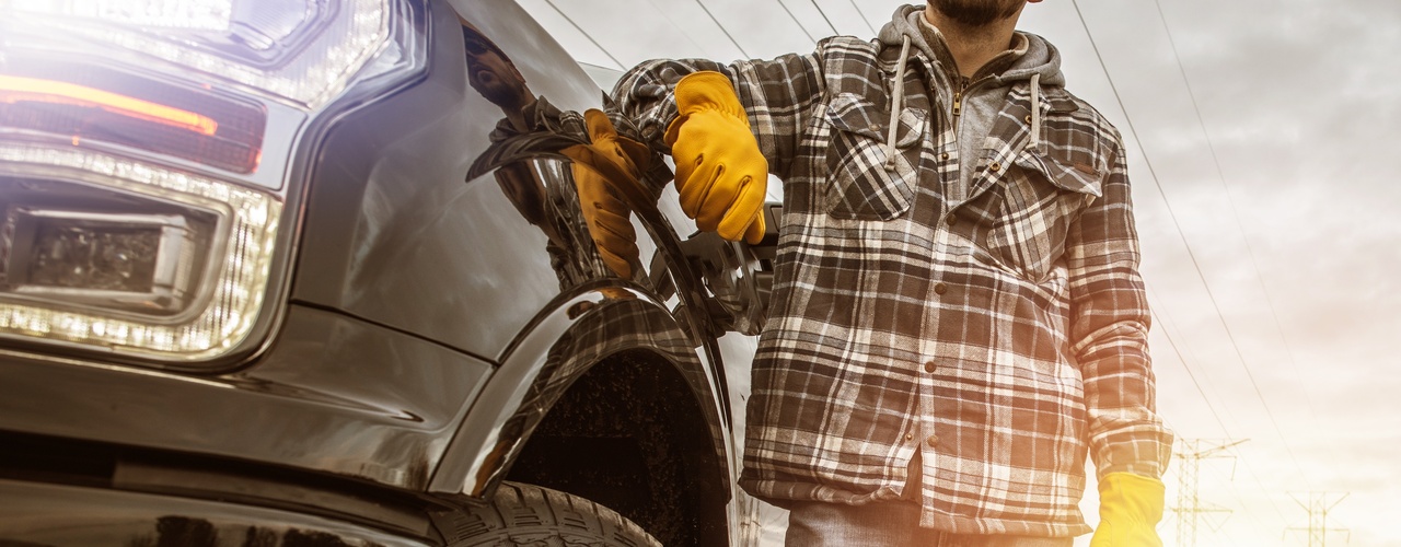 A man wearing a plaid shirt, yellow gloves and a helmet, standing next to a black pickup truck with the headlamps on.