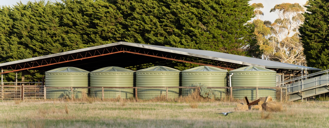 A view of five green water tanks stored near a covered storage shelter on a farm field with trees in the background.