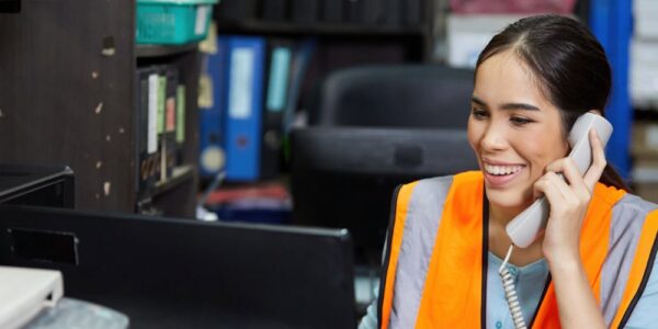 A woman in an orange safety vest smiling while holding a phone and looking at a computer monitor in an office.