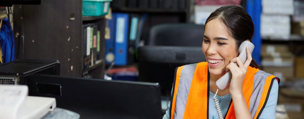 A woman in an orange safety vest smiling while holding a phone and looking at a computer monitor in an office.