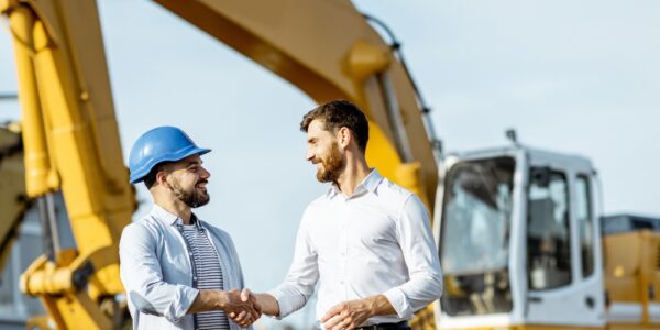 Two men shaking hands while standing in front of a piece of heavy construction machinery at an outdoor sales lot.