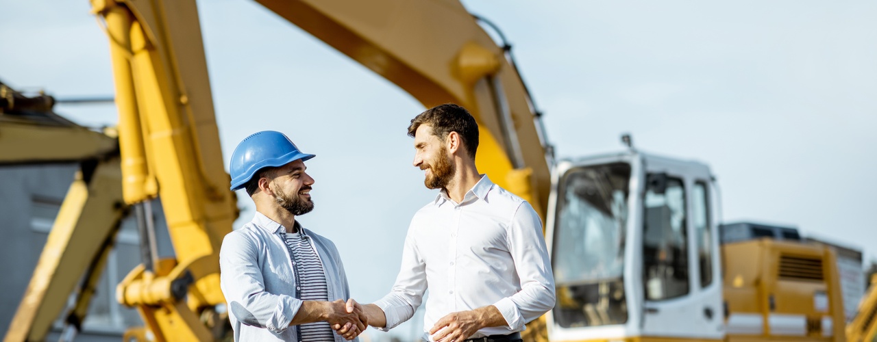 Two men shaking hands while standing in front of a piece of heavy construction machinery at an outdoor sales lot.