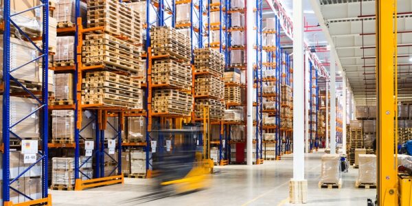 A warehouse facility lined with tall storage racks, boxes, overhead industrial lights, and forklifts going back and forth.
