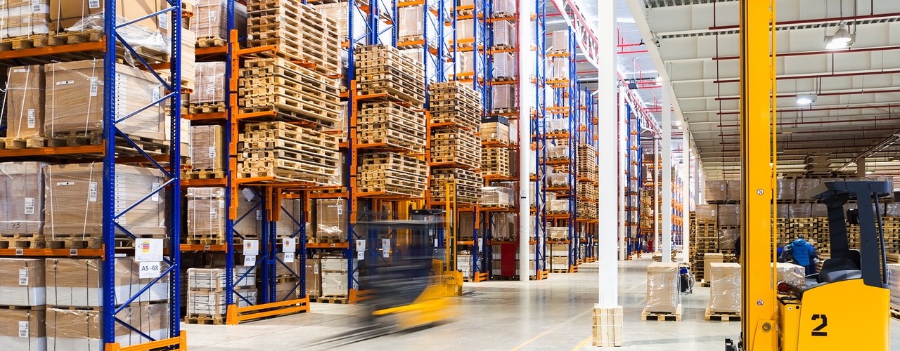 A warehouse facility lined with tall storage racks, boxes, overhead industrial lights, and forklifts going back and forth.