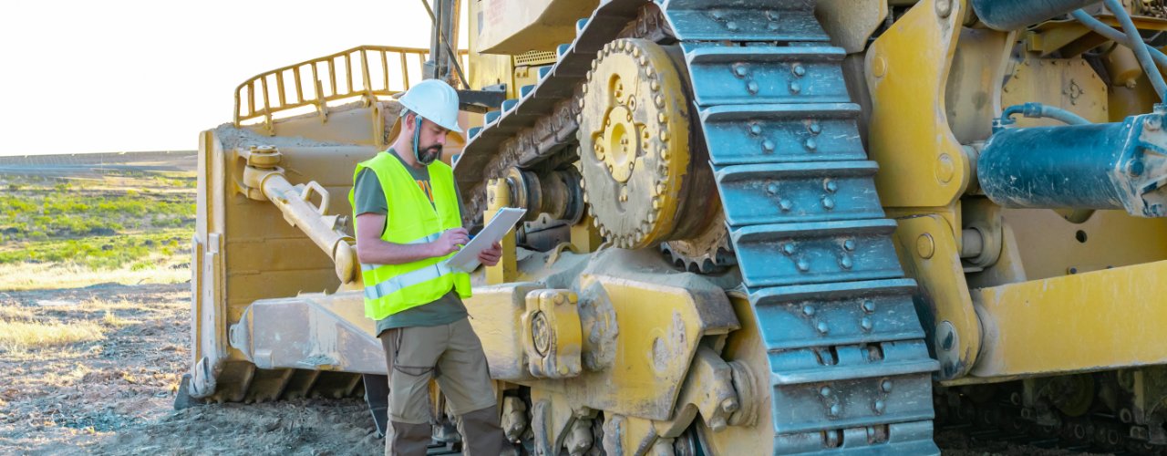 A man wearing a yellow reflective vest and a white hard hat stands at the base of a bulldozer. He takes notes on a clipboard.