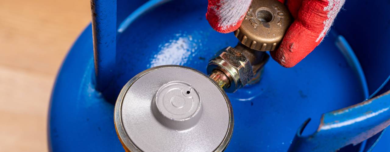 A close-up view shows a person wearing a work glove turning the bronze valve on a blue gas cylinder.