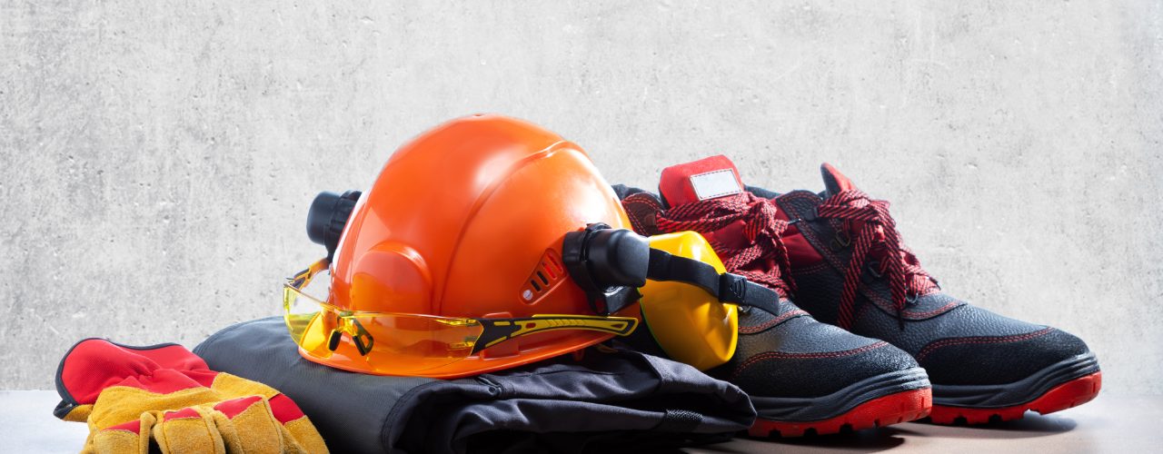A pile of work gear sits on the floor against a gray, concrete surface. There is a hat, gloves, and shoes.
