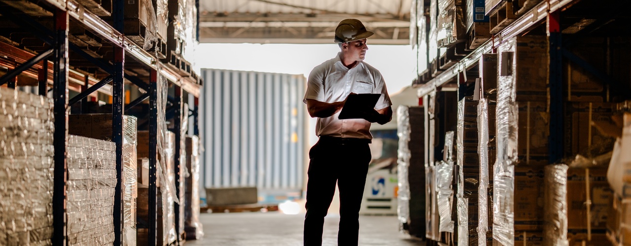 A man wearing a hard hat and holding a clipboard stands in the aisle of a warehouse. He looks at a pallet of supplies.