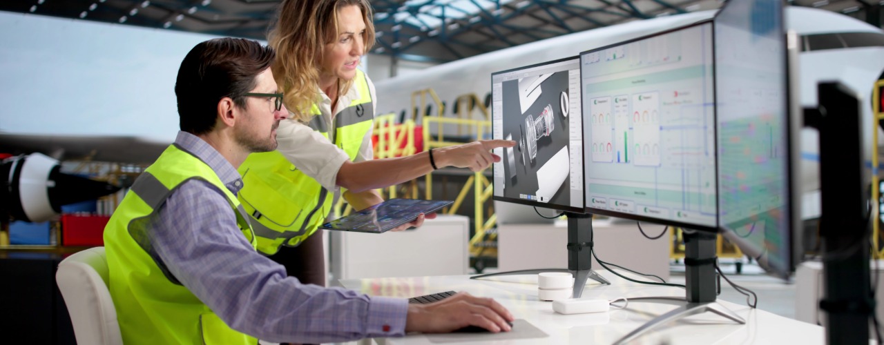 Two engineers convene at a desk. One sits behind three monitors while the other points at something on the screen.