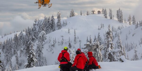 Five people kneel on a snowy mountaintop wearing bright red jackets and helmets. An orange helicopter is in the air.