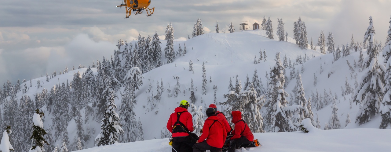 Five people kneel on a snowy mountaintop wearing bright red jackets and helmets. An orange helicopter is in the air.