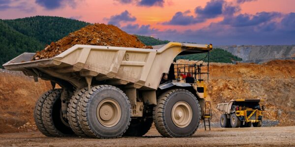 Large dump truck loaded with soil at an open-pit mine, with another truck and colorful sunset clouds behind.