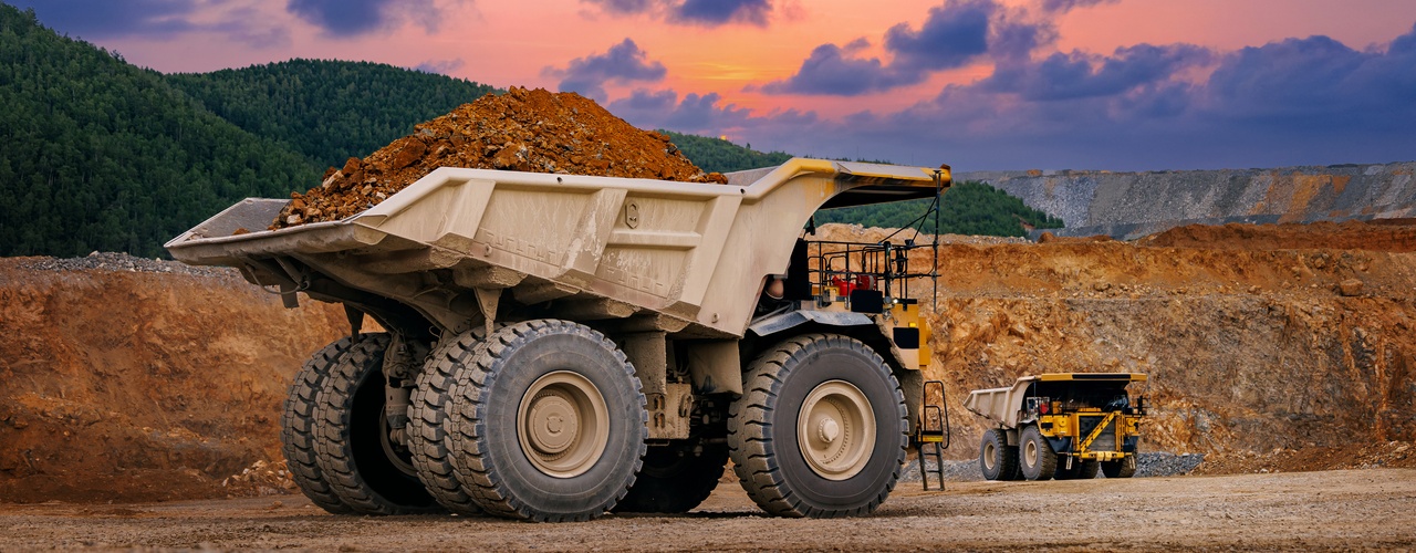 Large dump truck loaded with soil at an open-pit mine, with another truck and colorful sunset clouds behind.