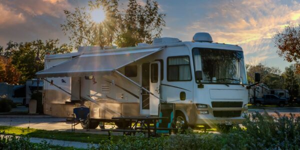 An RV parked at a campground at sunset, with an extended awning, picnic table, and trees glowing in evening light.