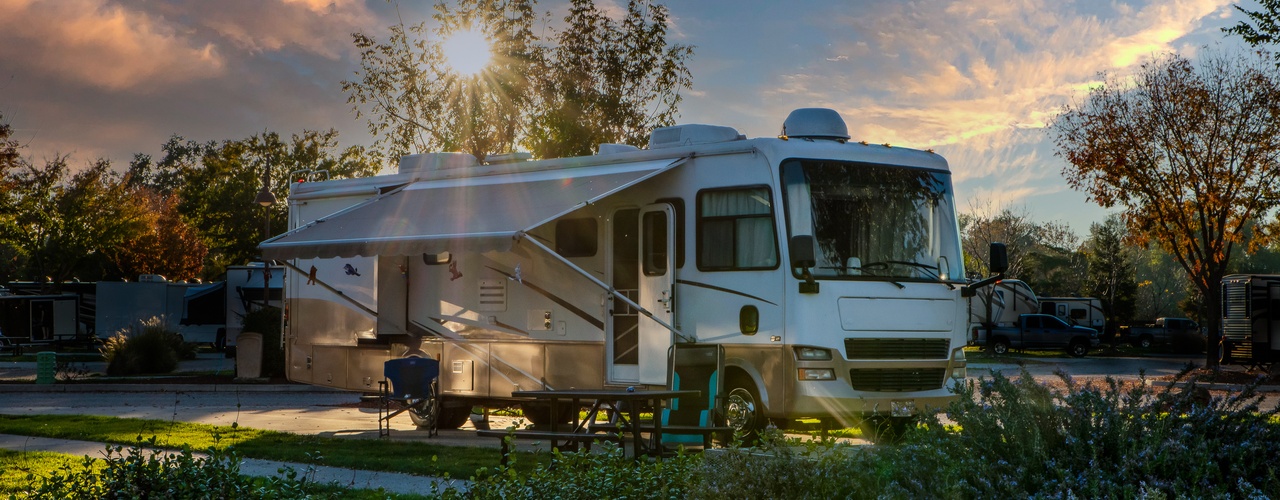 An RV parked at a campground at sunset, with an extended awning, picnic table, and trees glowing in evening light.