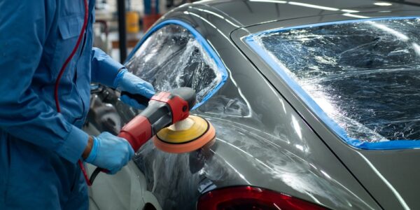 A person wearing a protective blue suit is using an electric buffer to polish a dark gray sports car.