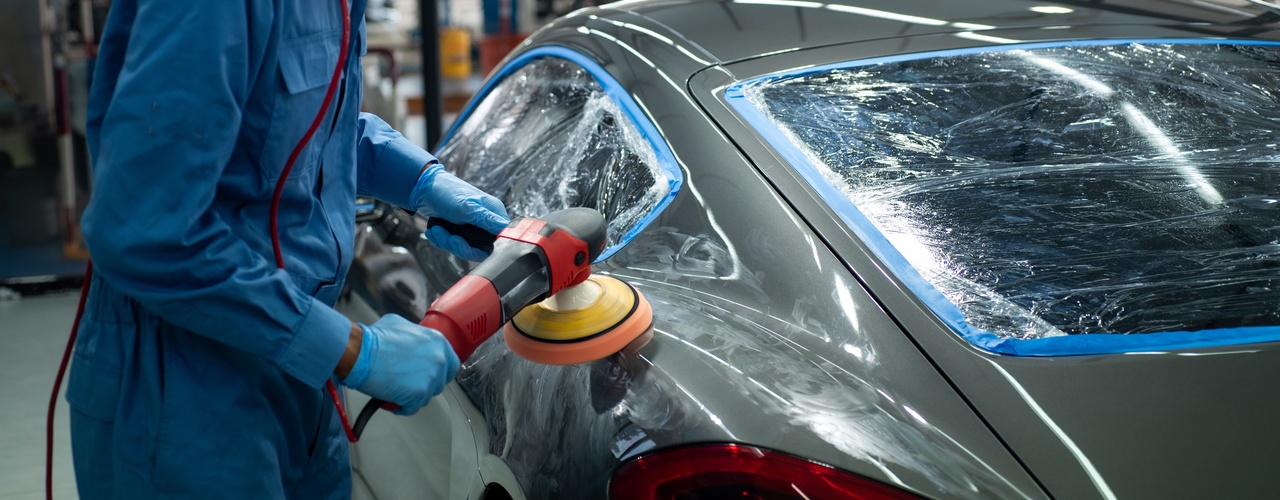 A person wearing a protective blue suit is using an electric buffer to polish a dark gray sports car.