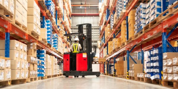 A wide warehouse aisle lined with tall shelves of boxed pallets as a worker drives a red forklift down the center.