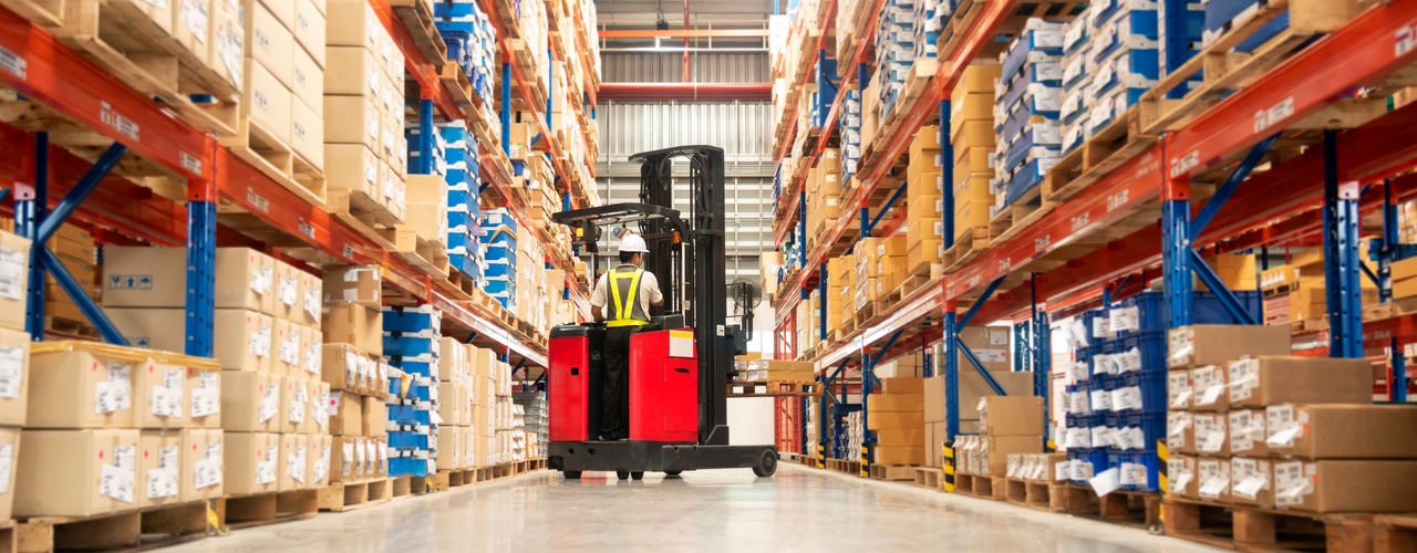 A wide warehouse aisle lined with tall shelves of boxed pallets as a worker drives a red forklift down the center.