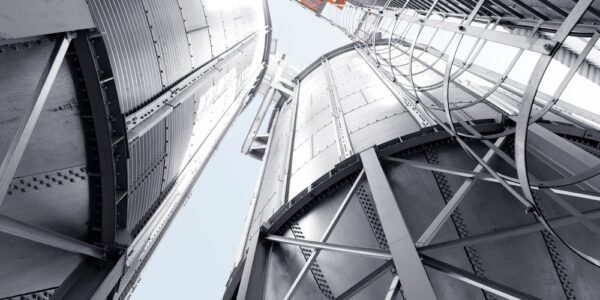 A skyward view between several tall metal silos with steel framework, ladders, and pale sky visible above the structures.
