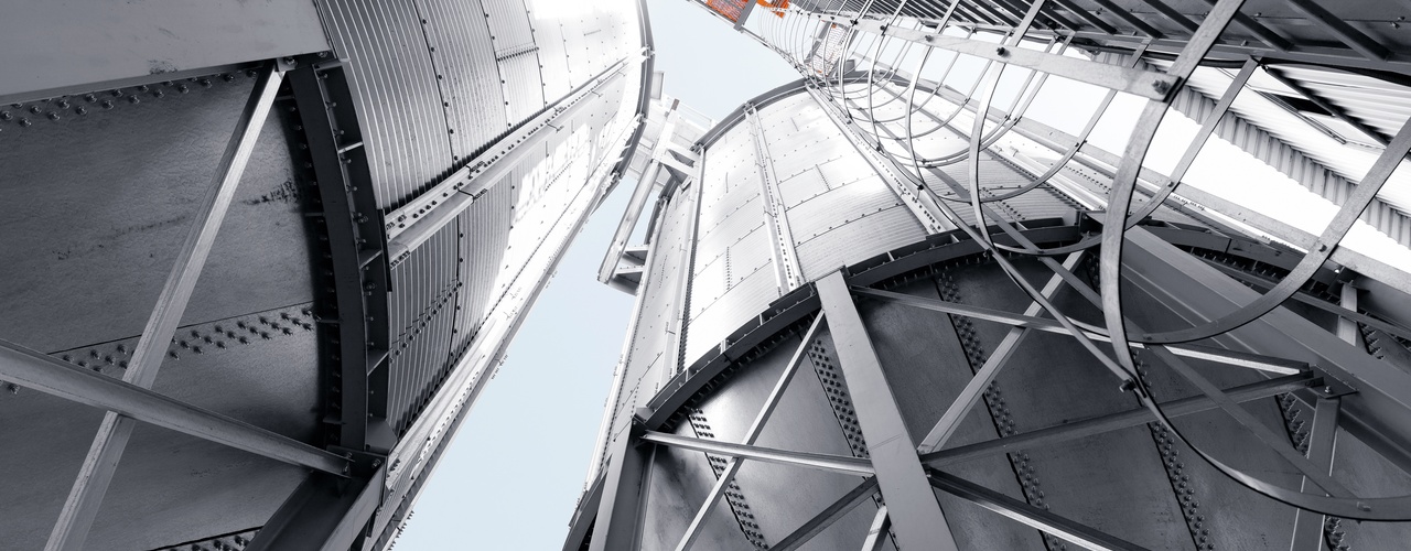 A skyward view between several tall metal silos with steel framework, ladders, and pale sky visible above the structures.