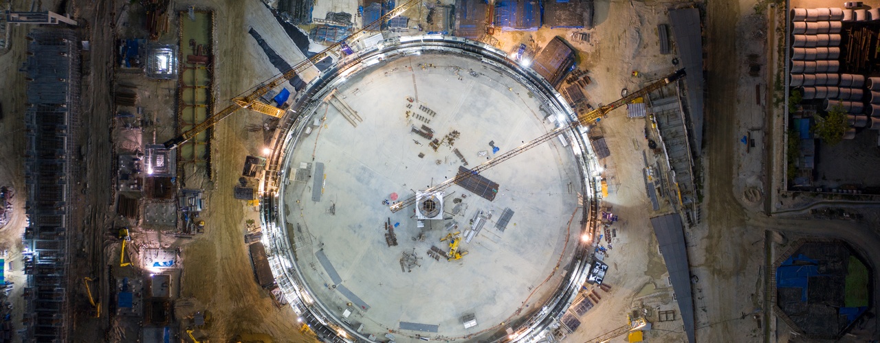 an illuminated industrial construction site at night with cranes, a circular structure, and surrounding work areas.