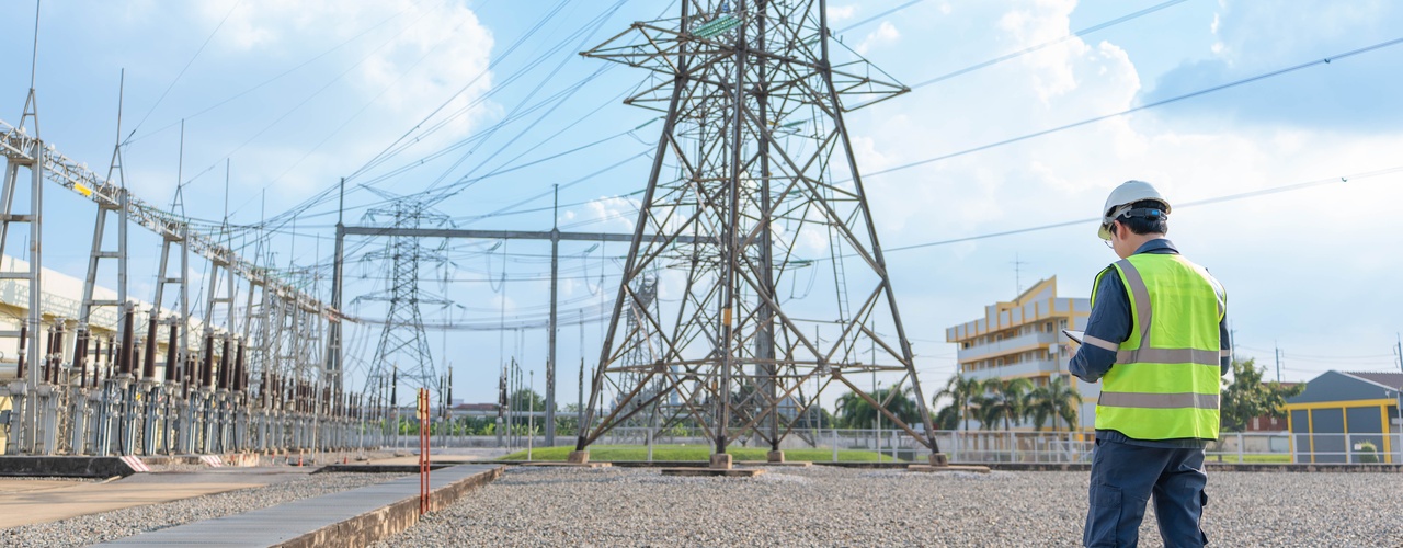 A man wearing a yellow safety vest and a white hard hat stands on a path in front of a transmission tower.