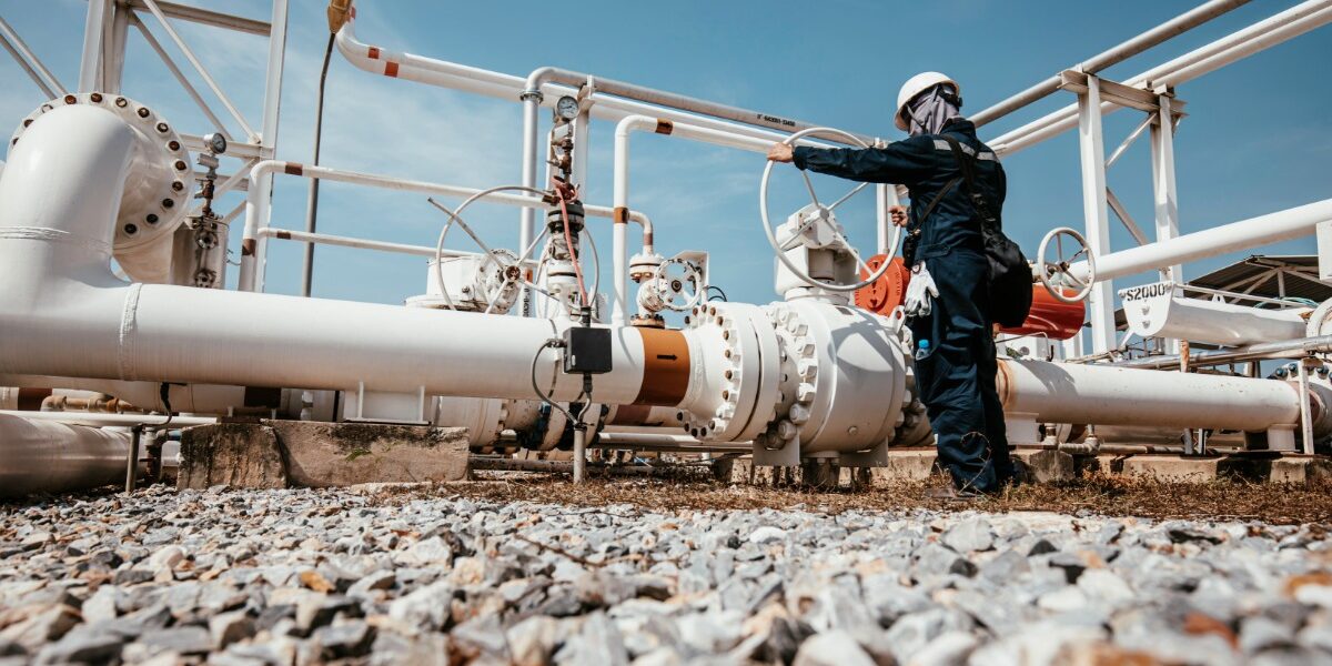 A worker wearing a hard hat operates a valve among white pipelines and metal equipment under a blue sky.