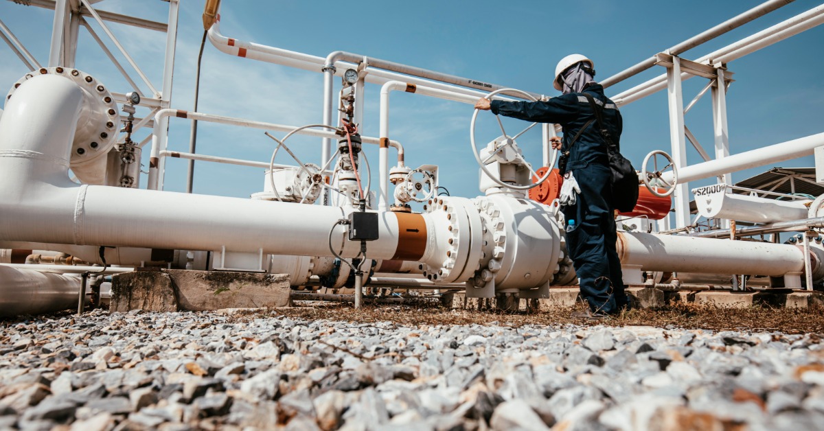 A worker wearing a hard hat operates a valve among white pipelines and metal equipment under a blue sky.