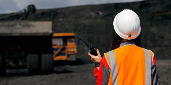 A worker in an orange reflective safety vest and white hard hat using a radio near a large dump truck at a mine site.