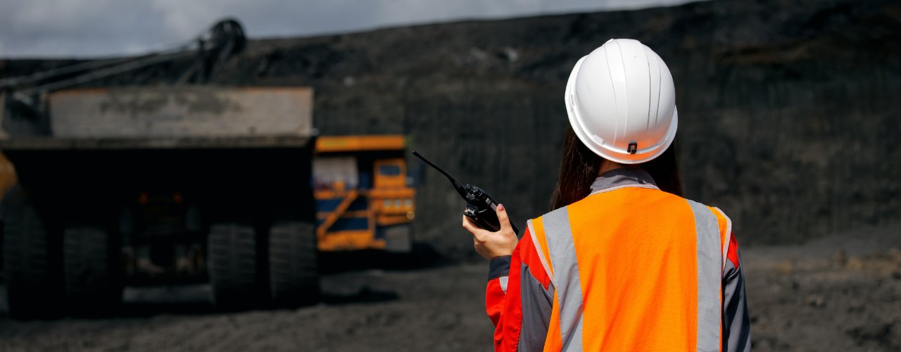 A worker in an orange reflective safety vest and white hard hat using a radio near a large dump truck at a mine site.