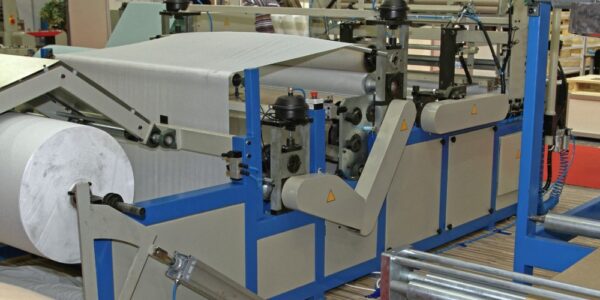 A large gray and blue industrial machine with robotic arms is rolling paper onto a tube from a belt.